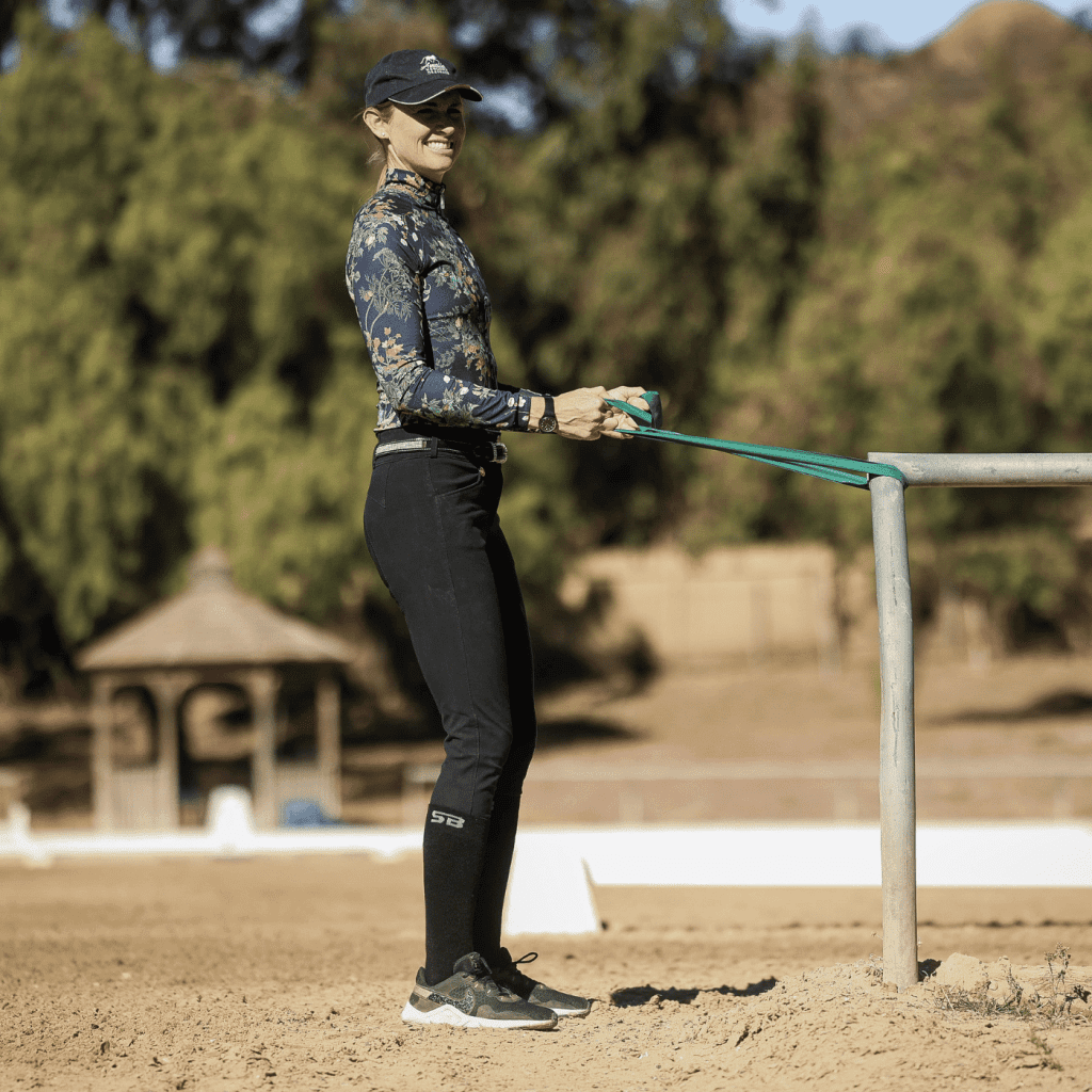 A woman in athletic wear demonstrates a Rider Position as she stretches a green resistance band attached to a metal rail outdoors on a sandy surface, with trees and a gazebo in the background.