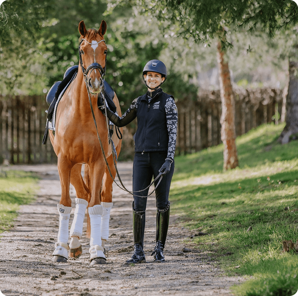A smiling person in riding gear, demonstrating the Rider Position Blueprint, leads a chestnut horse with white leg wraps along a sunlit, tree-lined path in a grassy area. Both appear calm and content.