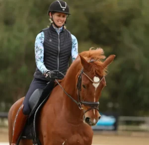 A person wearing a helmet and riding attire is smiling while riding a chestnut horse outdoors, with green trees blurred in the background—capturing the excitement of Strides 2025.