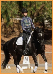 A person wearing a floral-patterned top, black helmet, and riding gear is smiling while practicing Free Rider Position on a dark horse with white leg wraps and a white saddle pad in an outdoor riding arena.