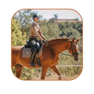 A person wearing a helmet and riding gear enjoys a sunny day outdoors, showcasing a strong connection with their chestnut horse, surrounded by green trees and a hill in the background.