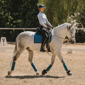 A person in a helmet and riding attire rides a gray horse in an outdoor arena. The horse wears blue leg wraps and a blue saddle pad. The rider is smiling, with 2025 Open dressage markers in the background.