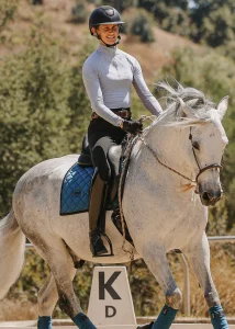 A person wearing a helmet and riding gear is smiling while riding a white horse with a blue saddle pad outdoors near a K dressage marker, preparing for the 2025 Open. Trees and grassy hills provide a scenic backdrop.