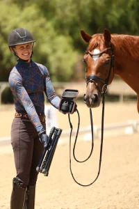 A smiling equestrian in riding gear and helmet holds a folded tripod and PIVO camera while standing beside a brown horse on a sandy outdoor arena with trees in the background. This popular item is currently Sold Out.