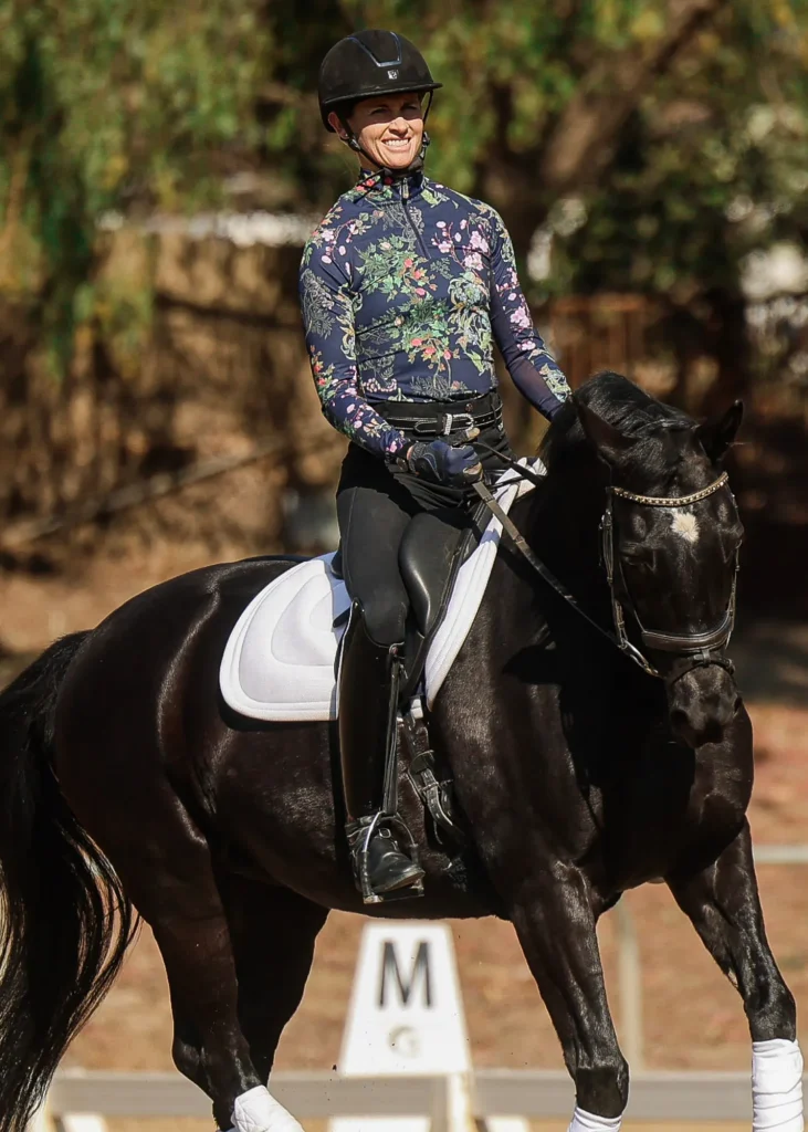 A person in a floral-patterned top, helmet, and riding boots smiles while riding a black horse with a white saddle pad during an OA Mini Course, showcasing their Free Rider Position. Trees and a dressage marker labeled M are visible in the background.