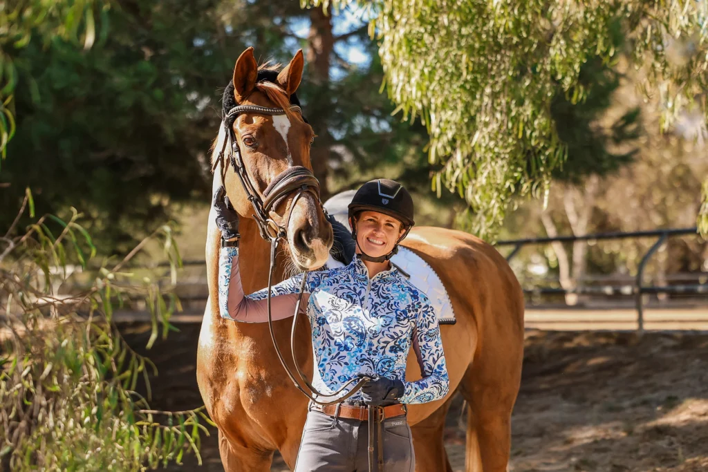 A person wearing a helmet and patterned shirt stands smiling next to a brown horse, holding its reins, in a sunlit outdoor setting with trees in the background.