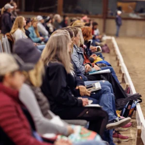 A group of people sit in rows of chairs indoors, attentively watching something off-frame. Many hold notebooks or papers, suggesting they are taking notes at a Grassroots Giving seminar or event.