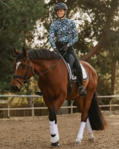 Amelia Newcomb, in helmet and riding attire, schools a bay horse with white markings and polos in an outdoor arena lined by trees.