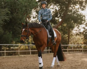 A person wearing a patterned long-sleeve shirt, helmet, and riding boots smiles while riding a brown horse with white leg wraps in an outdoor arena surrounded by trees—perfect for practicing Free Dressage Terms.