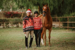 Two people in festive sweaters and riding boots stand beside a brown horse in a grassy, fenced area, celebrating the holiday spirit. One wears a Santa hat, the other a riding helmet. Trees fill the background. Perfect for holiday workshops or gatherings.