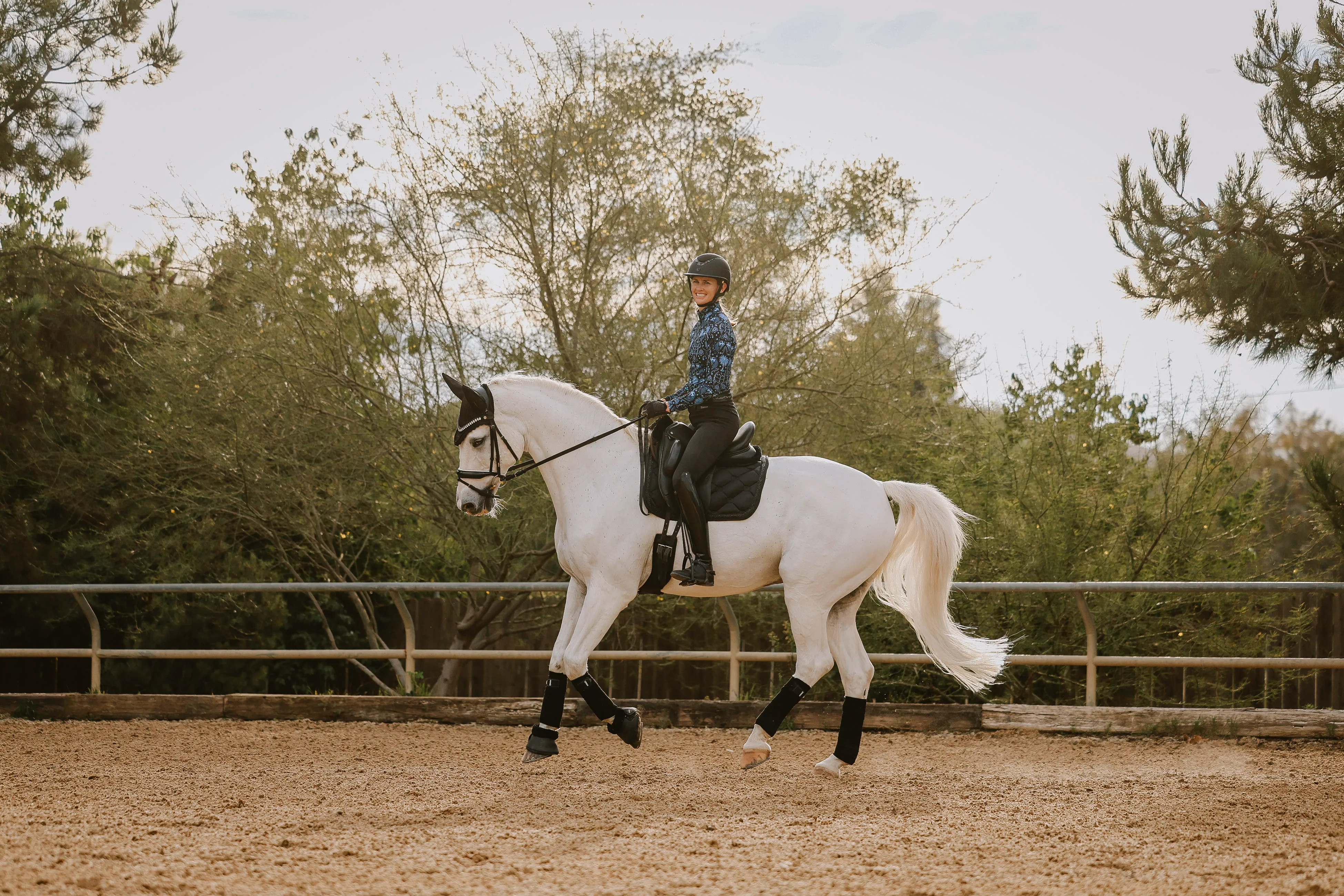 A person wearing a helmet and riding gear is engaged in dressage training, riding a white horse in an outdoor sandy arena surrounded by green trees and a wooden fence.