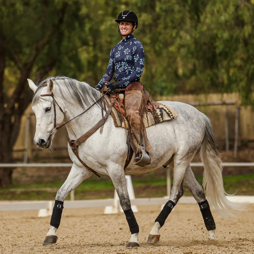 A person wearing a helmet and patterned shirt rides a light gray horse in an outdoor arena, practicing dressage foundations. The horse walks calmly, and both the rider and horse appear focused. Trees and a fence are visible in the background.