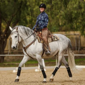 A person wearing a helmet and patterned shirt rides a light gray horse in an outdoor arena, practicing dressage foundations. The horse walks calmly, and both the rider and horse appear focused. Trees and a fence are visible in the background.