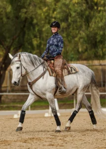 A person wearing a helmet, floral-patterned long-sleeve shirt, and brown chaps rides a gray horse in an outdoor sandy arena—an ideal setting for Dressage Foundations—surrounded by greenery and wooden fencing.