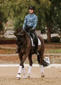 A smiling equestrian in a blue patterned shirt and black helmet rides a dark brown horse with white leg wraps in an outdoor arena, showcasing Dressage Foundations amid trees and fencing.