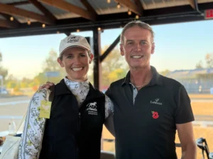 Amelia in a branded vest and Carl Hester in a black polo shirt stand side by side, smiling for a photo.