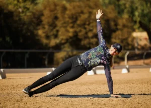 A person wearing a hat, sunglasses, and athletic clothing performs a side plank on a mat outdoors on a sandy surface, enjoying an active holiday with trees and a fence in the background.