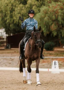 A smiling equestrian in a blue patterned shirt, black helmet, gloves, and boots waves while riding a dark horse with white leg wraps—showcasing Dressage Foundations—on a sandy arena with green trees and a fence in the background.