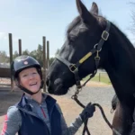 A smiling person in a riding helmet stands next to a large black horse, holding its reins outdoors on a sunny day. The horses head is close to the person, creating a friendly and joyful moment.