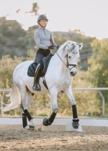 A person in riding gear and a helmet is riding a white horse in an outdoor arena, demonstrating the Training Scale. The horse is trotting with focus, while trees and a fence form the backdrop—a scene fit for a Scale Masterclass.
