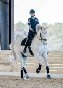 A person wearing a helmet, vest, and patterned sleeves rides a white horse inside an arena with wooden walls and sandy ground. Both look focused, embodying Dressage Foundations as they train for the Early Bird event in July 2026.