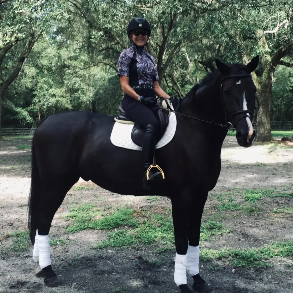 A rider in a helmet and breeches is mounted on a black horse (possible black or dark bay) with white polos and dressage pad in an outdoor wooded arena.