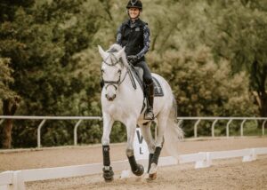 A person wearing a helmet and dark riding gear is smiling while riding a white horse in an outdoor arena, surrounded by trees and a fence—perfect for equestrian masterclasses.