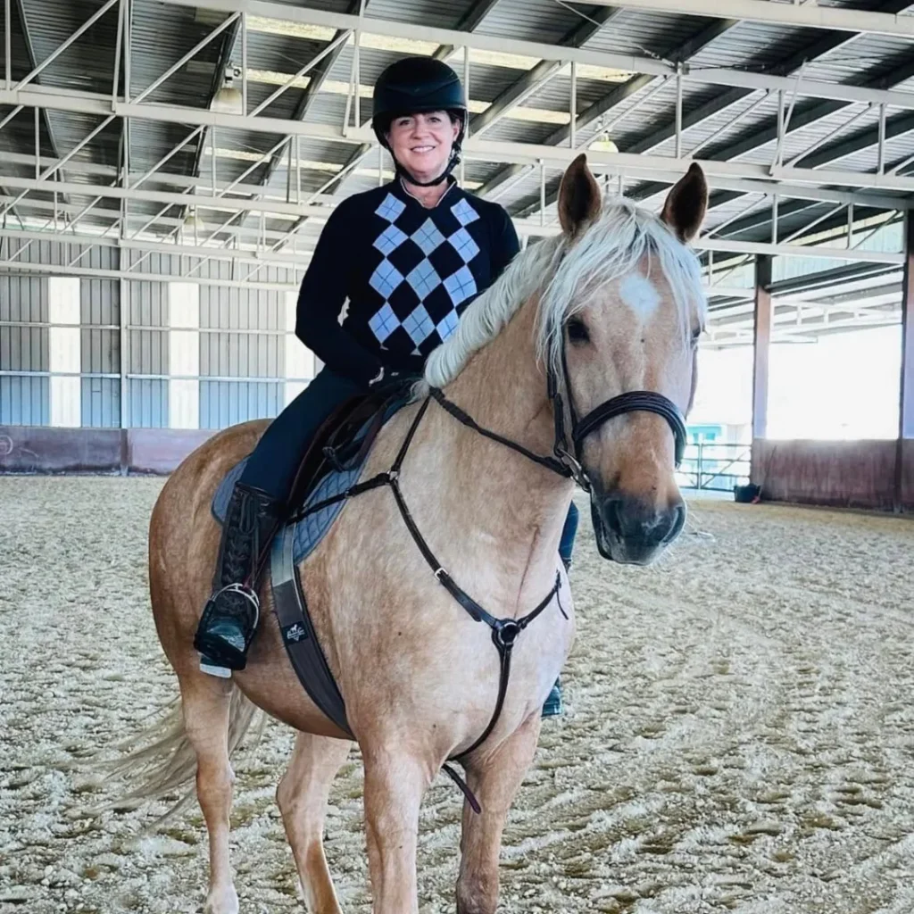 A rider in casual attire sits on a palomino horse in an indoor arena with sand footing, prepared for flatwork or basic schooling.