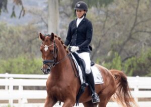 A person in formal equestrian attire rides a brown horse during a dressage event outdoors, with white fences and trees in the background, showcasing skills often refined through masterclasses.