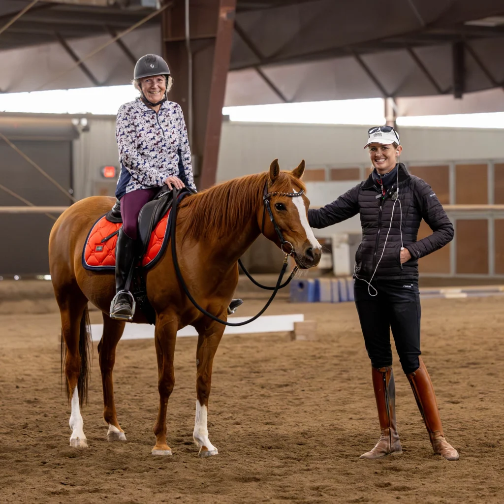 A student in casual attire and helmet sits on a chestnut gelding with an orange saddle pad as a coach observes in the indoor arena.