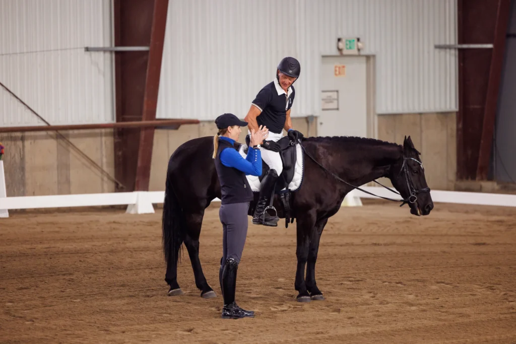 A person riding a black horse inside an indoor arena reaches down to high-five instructor Amelia Newcomb, both wearing helmets and riding gear. The arena features brown dirt and white fences.