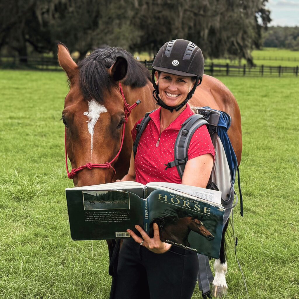 A student in a helmet and red shirt stands on grass, holding an open equestrian training manual. A bay horse with a distinct white blaze gently nuzzles her and the book, demonstrating a relaxed connection consistent with groundwork and horsemanship principles. Behind them, trees and a rail fence provide the classic backdrop of an outdoor riding arena, reflecting the focus on foundational skills and horse-human partnership typical of Riding Bootcamp sessions.