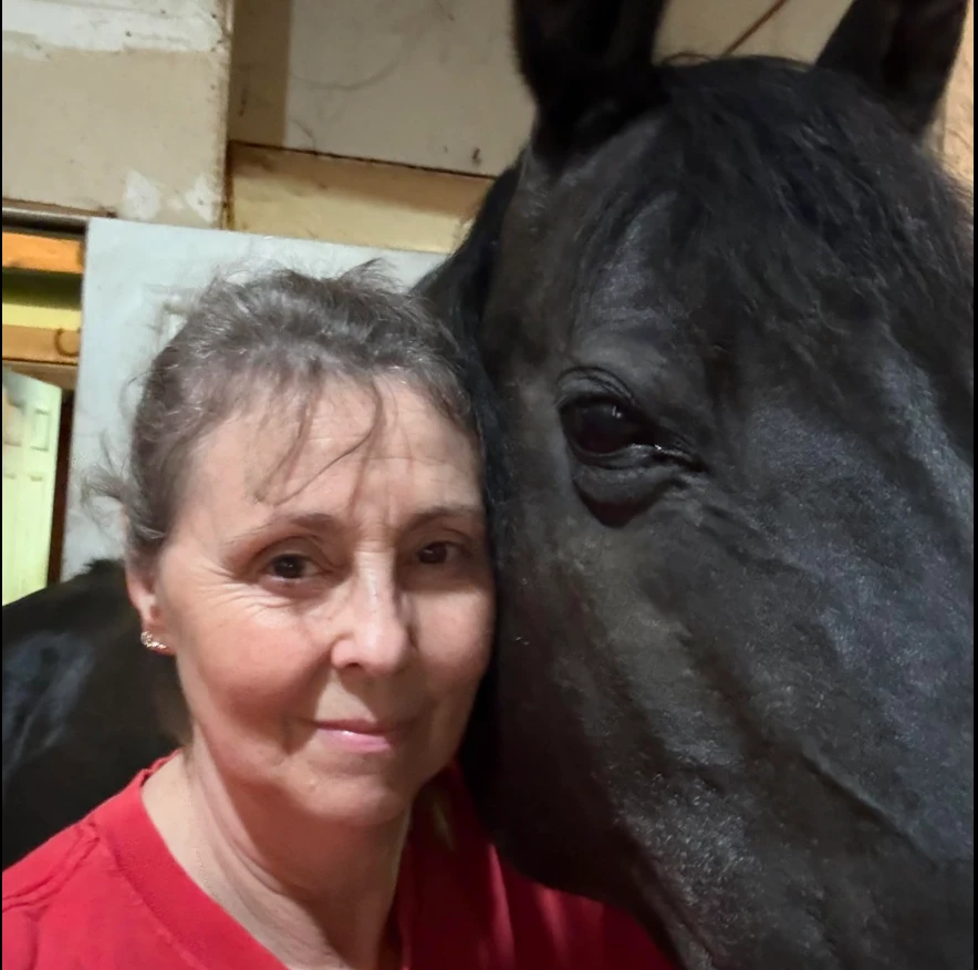 A rider with grey hair and a red shirt stands beside a large black horse, both facing the camera in an indoor arena setting, positioned as if for a testimonial photo. The horse’s coat is true black without markings, and their relaxed demeanor suggests they are between dressage schooling movements.