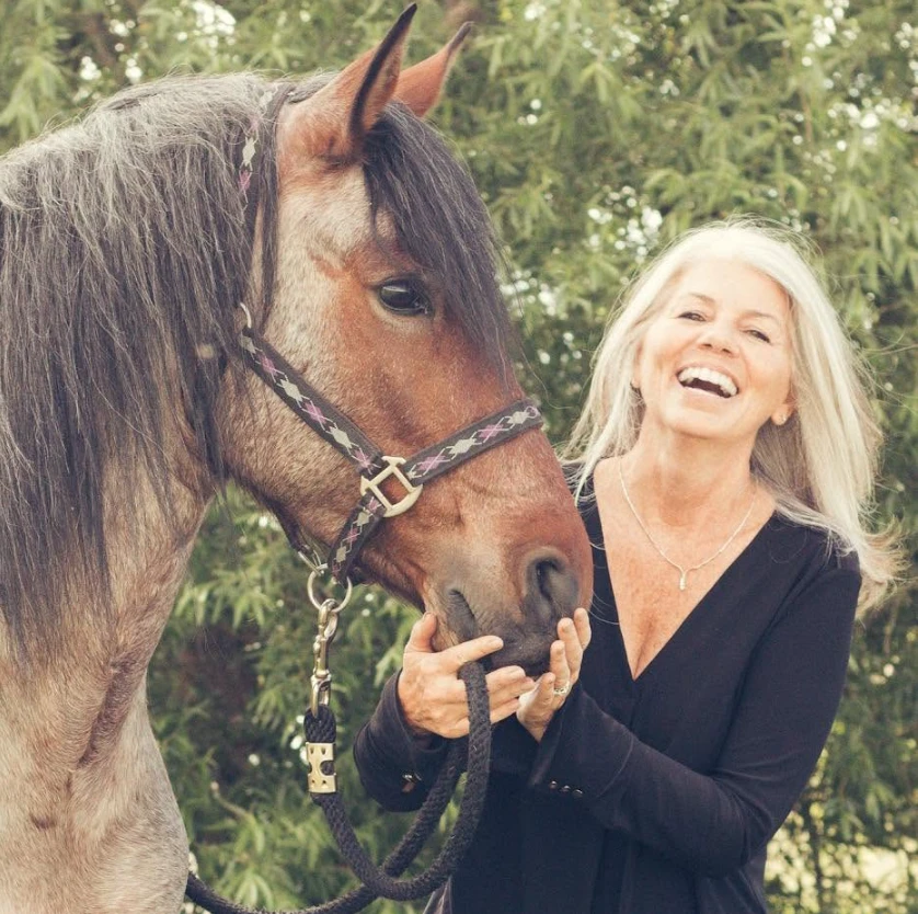 A smiling rider with long gray hair stands outdoors beside a bay horse, gently holding its halter in a relaxed stance reminiscent of a schooling break, as if ready to share her story for a Testimonial Slider. Lush green foliage creates a natural background.