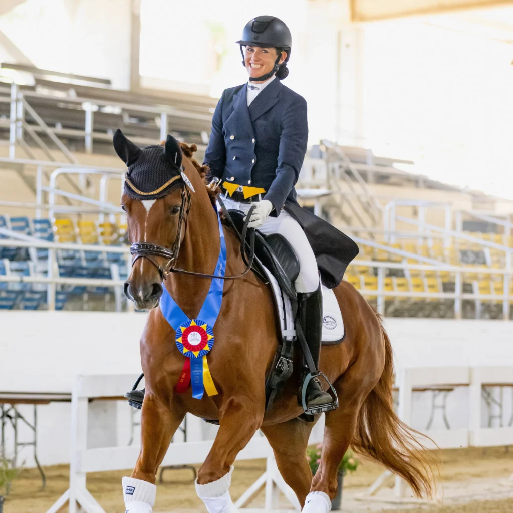Amelia Newcomb, dressed in professional dressage attire with a shadbelly and top hat, smiles while riding a bay horse performing an extended trot in an indoor arena. The horse proudly wears a blue, yellow, and red winner’s sash draped around its neck, celebrating their victory at the 09/2026 competition highlighted on the Opt-In Page.