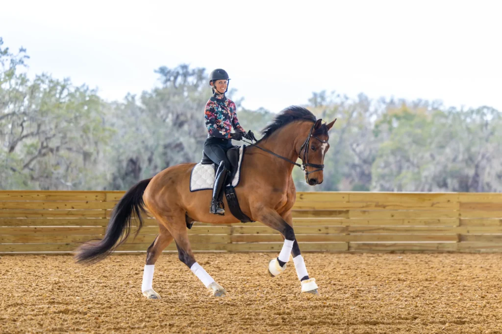 A student performs an extended trot on a bay horse while wearing a floral shirt in a sandy dressage arena.