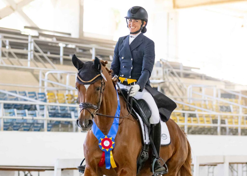 Amelia Newcomb, attired in professional dressage clothing including a shadbelly coat, stock tie, and white breeches, sits confidently atop a bay horse displaying a blue and yellow championship rosette on its bridle. They are positioned inside an indoor arena with vacant spectator stands and signage promoting Free Resources visible in the background.