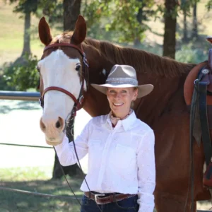A smiling woman in a white shirt and tan hat stands beside a brown horse with a white face, holding its bridle outdoors on a sunny day. Trees and a fence are visible in the background.