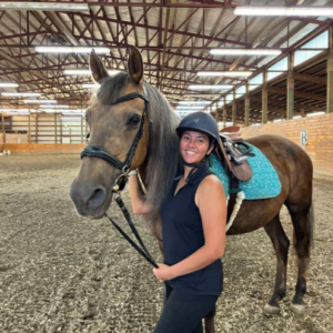 A smiling woman in a black helmet and riding attire stands next to a brown horse with a teal saddle pad inside an indoor riding arena with a high ceiling and exposed beams.
