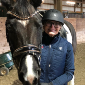 A person wearing glasses, a helmet, and a blue vest stands next to a black and white horse inside an indoor riding arena with wooden walls, ready for the Flying Changes Webinar.