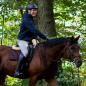 A person wearing a black helmet, dark jacket, and light pants rides a brown horse through a wooded area with green foliage, practicing skills learned from the Flying Changes Webinar.