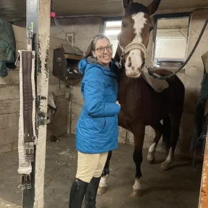 A woman in a blue jacket and beige pants stands smiling next to a brown horse with a white stripe on its face inside a stable. The horse is wearing a halter and a blanket over its back.