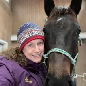 A smiling woman in a purple jacket and patterned winter hat stands next to a dark brown horse wearing a blue and white rope halter. They are indoors, close together, facing the camera.
