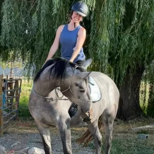 A smiling person wearing a helmet and blue sleeveless top sits on a gray horse, outdoors in front of a leafy green tree. The horse is turning its head to the left.