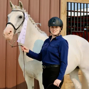 A person in a helmet and blue shirt stands beside a white horse with braided mane, holding a first-place ribbon. They are both in front of a brown barn wall and open stable door.