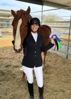 Esther Nava wearing show attire, smiling and holding a rosette while posing with her horse.
