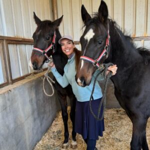 A smiling woman in a hat stands between two dark brown horses with red halters inside a stable, holding their lead ropes. The stable has a metal roof and wooden walls, and straw covers the floor.