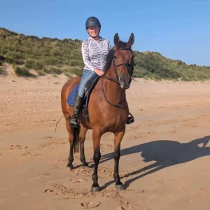 A person wearing a helmet and patterned shirt smiles while sitting on a brown horse on a sandy beach with grassy dunes in the background under a clear blue sky.