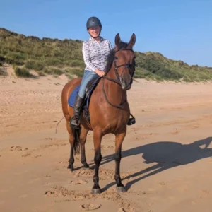 A person wearing a helmet and patterned shirt smiles while sitting on a brown horse on a sandy beach with grassy dunes in the background under a clear blue sky.