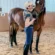 A rider in casual attire stands in an indoor arena, gently holding and kissing the face of a bay horse.